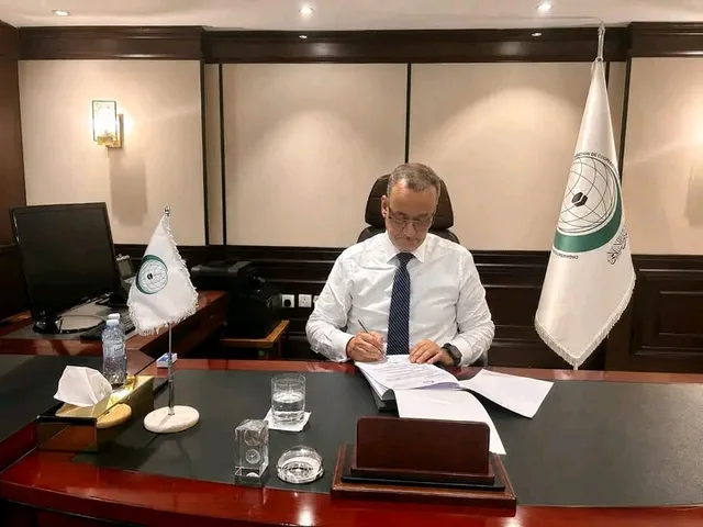 Le Secrétaire général de l'Organisation de la coopération islamique signe des documents à son bureau, près des drapeaux de l'organisation