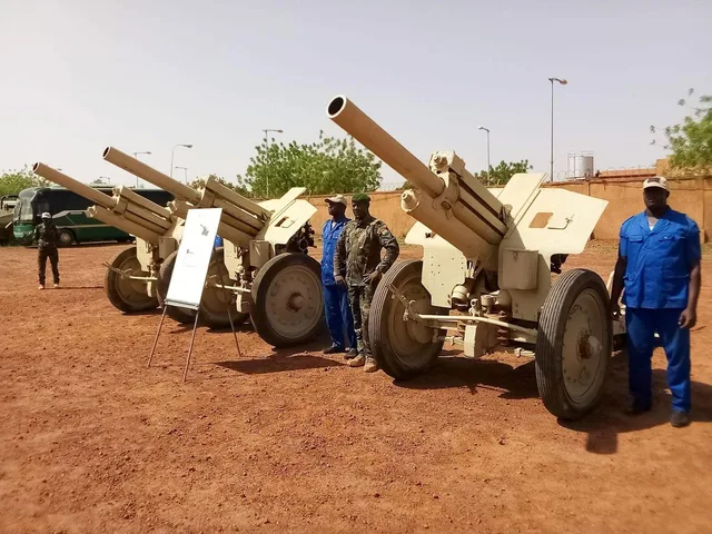 Trois canons de campagne sur un sol de latérite, à côté d'un militaire et d'agents en tenue bleue, photo d'archives