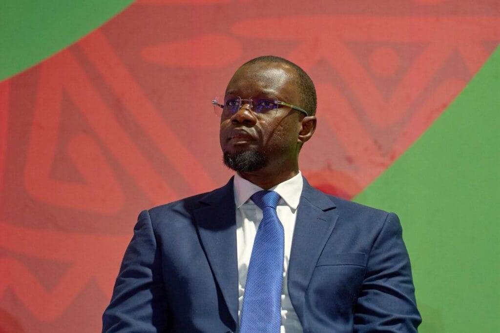 Ousmane Sonko en costume bleu devant un fond aux couleurs rouge et verte