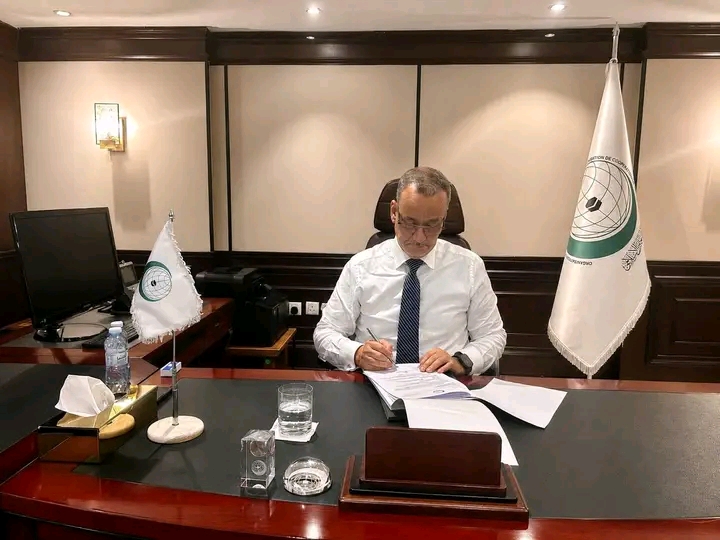 Le Secrétaire général de l'Organisation de la coopération islamique signe des documents à son bureau, près des drapeaux de l'organisation